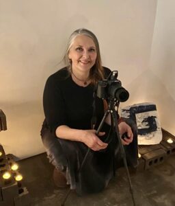 Helen smiling as she kneels beside a camera on a tripod, dressed in black with a softly lit background and art pieces nearby.