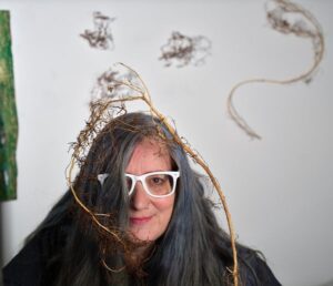 Headshot of Lauri with long gray hair and white-framed glasses, adorned with natural twigs, against a white background.