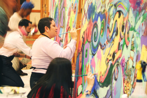 A group of people stand painting in front of the large colorful mural they are working on