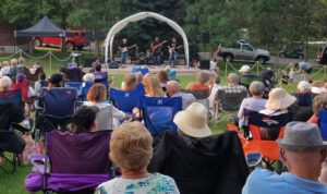 A group of people sitting on a lawn listening to a band perform on a small stage.