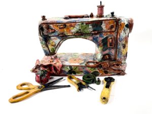 A ceramic sewing machine sculpture that is brightly painted. Ceramic sculptures of sewing notions including a pair of scissors, pin cushion, seam ripper, tape measure, and rotary cutter lay in front of the sewing machine